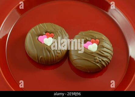 Two Valentine`s Day chocolate dipped sandwich cookies with heart decorations on red plate on pink background Stock Photo